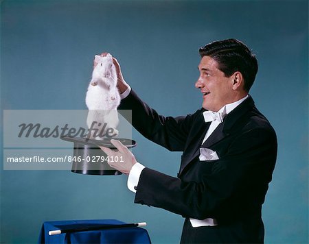 1960s MAGICIAN WEARING BLACK TUXEDO SMILING PULLING WHITE RABBIT OUT OF TOP HAT