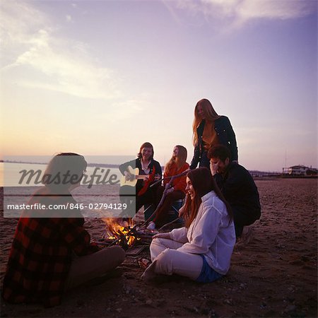 1970s GROUP TEENAGER BOYS GIRLS TOGETHER SEASHORE SHORE CAMPFIRE GUITAR SINGING RETRO SAND SUNSET OCEAN BEACH OUTDOOR