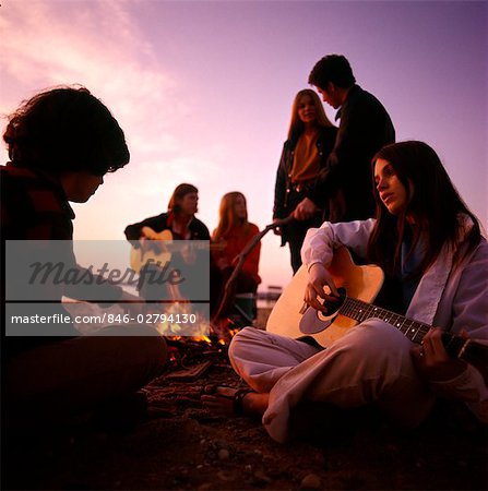 1970s TEENAGERS GATHERED AROUND BEACH CAMPFIRE PLAYING ACOUSTIC GUITARS
