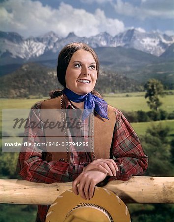 1960s WOMAN IN WESTERN OUTFIT LEANING ON RAIL FENCE HOLDING HAT MOUNTAIN SCENE IN BACKGROUND STUDIO OUTDOOR