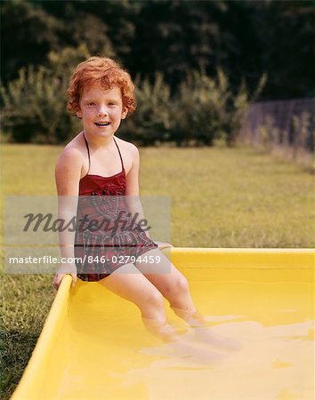 1960s SMILING RED HAIRED LITTLE GIRL IN PLAID BATHING SUIT SITTING ON YELLOW PLASTIC BACKYARD SWIMMING POOL