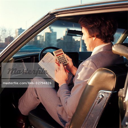 1970s MAN BUSINESSMAN SALESMAN USING POCKET CALCULATOR SITTING IN AUTOMOBILE