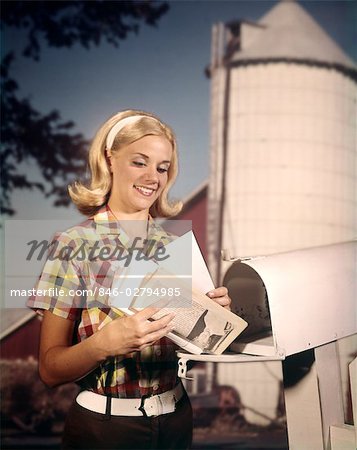 1960s SMILING YOUNG BLOND WOMAN TAKING MAIL FROM RURAL DELIVERY MAIL BOX MAILBOX FARM SILO BACKGROUND