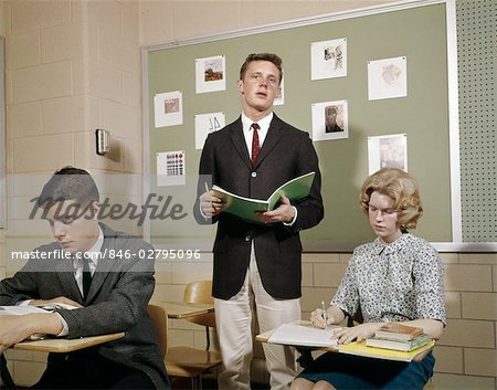 1960s TEEN STUDENTS BOYS GIRLS CLASSROOM BOY STANDING READING FROM NOTEBOOK HIGH SCHOOL