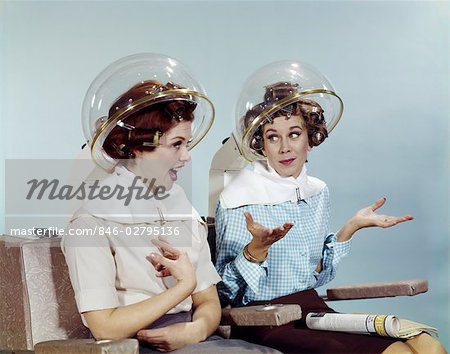 1960s TWO WOMEN SITTING UNDER BEAUTY SALON HAIR DRYER HOODS IN CURLERS TALKING GOSSIP