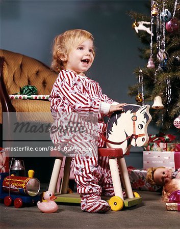 1960s TODDLER GIRL ON WHEELED TOY RIDING HORSE NEAR CHRISTMAS TREE LIVING ROOM HAPPY SMILING