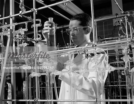 1960s MAN IN LAB COAT & GOGGLES SURROUNDED BY GLASS TUBING CONDUCTING SCIENTIFIC RESEARCH