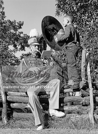 1950s MAN PLAYING A TUBA SITTING ON A LOG FENCE IN A BAND UNIFORM WHILE A BOY WHO IS STANDING ON THE FENCE HOLDING A FOOTBALL WATCHES