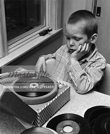 1960s 1970s BOY WITH BUZZ HAIRCUT CHIN IN HAND SITTING AT TABLE LISTENING TO MUSIC ON SMALL PORTABLE 45 RPM PHONOGRAPH RECORD PLAYER