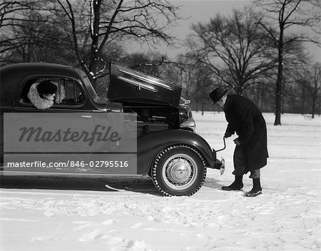 1930s 1940s WOMAN PASSENGER WATCHING MAN MOTORIST TRY TO CRANK START A CHEVROLET COUPE STALLED IN SNOW