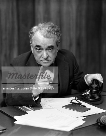 1930s HEAD-ON SERIOUS SENIOR BUSINESSMAN BEHIND DESK HAND ON TELEPHONE POINTING