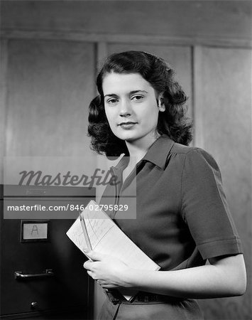 1940s 1950s WOMAN STANDING NEAR FILING CABINET HOLDING PEN AND PAPER