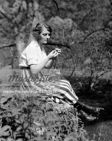 1920s YOUNG WOMAN IN STRIPED SKIRT SITTING ON STREAM BANK IN WOODS BRAIDING GARLAND OF WILD FLOWERS