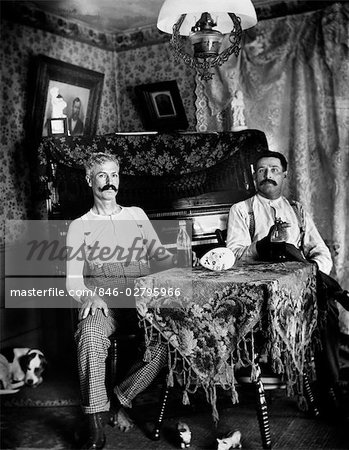 1890s TWO MEN IN SHIRT SLEEVES SITTING AT TABLE DRINKING BOTTLES OF BEER WITH PIANO AND DOG IN BACKGROUND