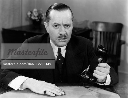 1930s 1940s PORTRAIT OF BUSINESS MAN SITTING AT OFFICE DESK HOLDING PHONE IN ONE HAND SERIOUS FACIAL EXPRESSION MOUSTACHE