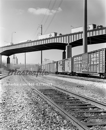 1970s ANGLED VIEW OF FREIGHT TRAIN PASSING UNDER HIGHWAY BRIDGE