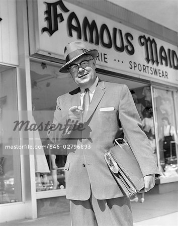 1950s MAN POINTING TO CAMERA HOLDING BRIEFCASE UNDER ARM IN FRONT OF SPORTS WEAR STORE SELL SUIT TIE HAT BUSINESSMAN