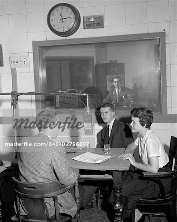 1960s DISC JOCKEY AT RADIO STATION MIKE MICROPHONE TALK WITH COUPLE MAN WOMAN AT TABLE MAN IN CONTROL ROOM