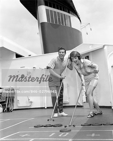 COUPLE MAN WOMAN SHIPBOARD PLAY PLAYING SHUFFLE BOARD ABOARD SHIP EXPRESSION FUN HAPPY VACATION SUMMER 1960s