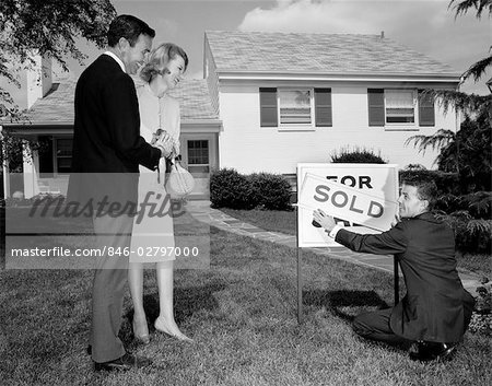 1960s SMILING COUPLE STANDING ON FRONT LAWN OF NEW HOUSE LOOKING DOWN AT REALTOR PUTTING UP SOLD SIGN