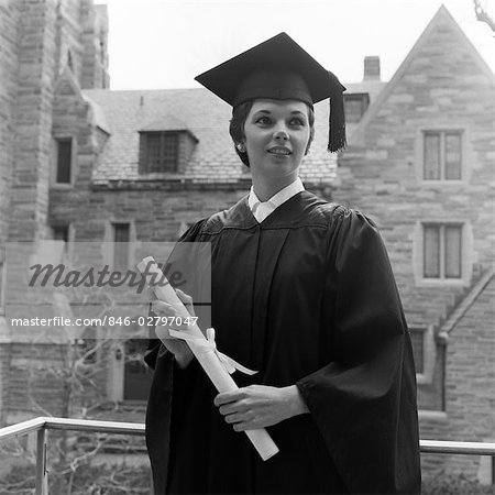 1950s SMILING FEMALE GRADUATE HOLDING DIPLOMA WITH STONE CAMPUS BUILDINGS IN BACKGROUND