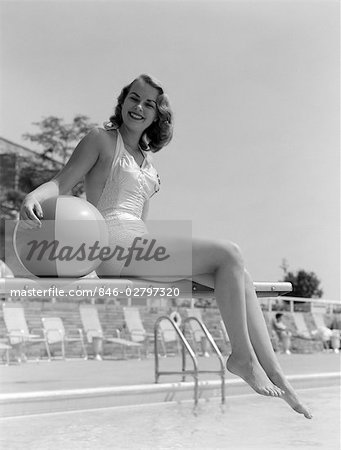1950s BATHING BEAUTY IN SATIN ONE-PIECE SITTING WITH LEGS HANGING OFF DIVING BOARD HOLDING BEACH BALL