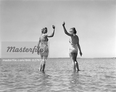 1930s TWO GIRLS STANDING IN WATER WEARING BATHING SUITS LOOKING BACK WAVING