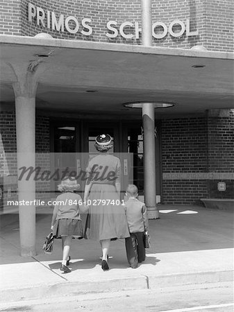 1950s BACK VIEW OF MOTHER HOLDING HANDS OF SON & DAUGHTER WALKING THEM INTO SCHOOL