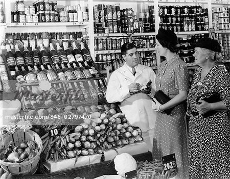 1940s GROCER IN WHITE COAT HOLDING PAD & PENCIL TO TAKE DOWN ORDERS FROM TWO WOMEN TALKING TO HIM