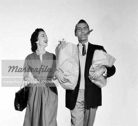 1950s COUPLE WALKING WOMAN SMILING AT MAN CARRYING GROCERY BAGS SMOKING CIGAR