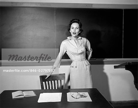 1950s SCHOOL TEACHER STANDING IN FRONT OF BOARD LEANING ON CHAIR BY DESK