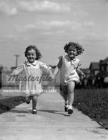1930s TWO CHILDREN RUNNING SKIPPING ON SIDEWALK SMILING