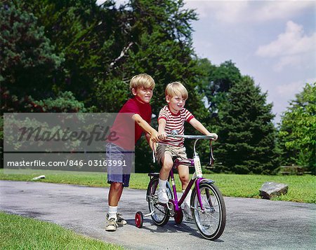 1960s 1970s BOY HELPING LITTLE BROTHER RIDE TWO WHEEL BICYCLE WITH TRAINING WHEELS