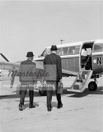 1960s TWO BUSINESSMEN WALKING TOGETHER TO A SMALL PRIVATE PROPELLER AIRPLANE CARRYING BRIEFCASE