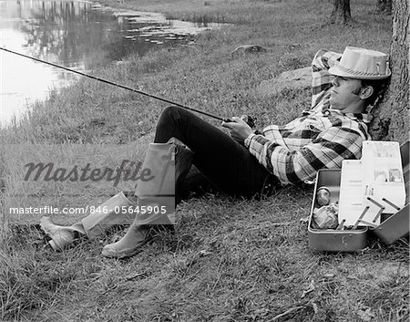 1970s MAN SLEEPING AGAINST A TREE TRUNK WHILE FISHING IN A LAKE