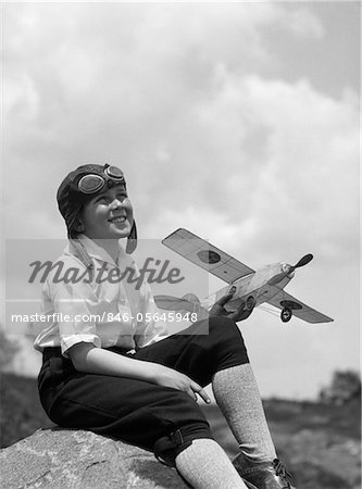 1930s BOY IN LEATHER AVIATOR CAP WITH GOGGLES SITTING ON ROCK HOLDING MODEL PLANE