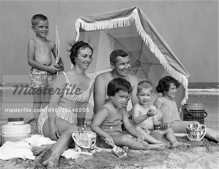 1960s FAMILY OF 6 ON BEACH UNDER UMBRELLA WITH PAILS AND COOLERS