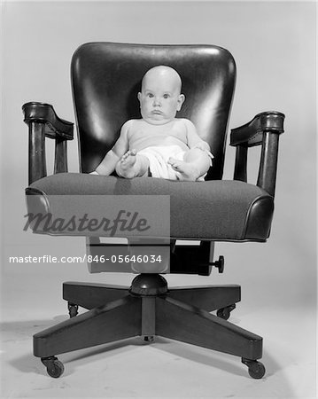 1960s PORTRAIT OF CHUBBY BALD BABY IN DIAPER SITTING IN EXECUTIVE OFFICE CHAIR