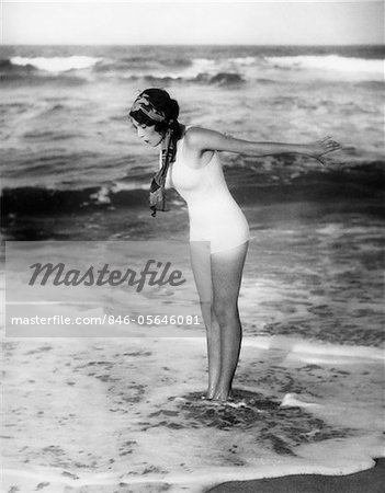 1920s WOMAN WEARING BATHING SUIT & HEAD SCARF LOOKING DOWN AT WATER STANDING IN OCEAN SURF