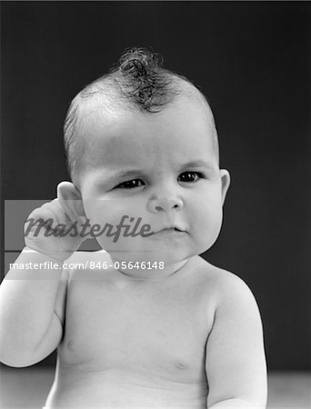 1940s BABY WITH SLIGHT SQUINTING EYES AND PUCKERED MOUTH HOLDING RIGHT HAND BEHIND RIGHT EAR STUDIO