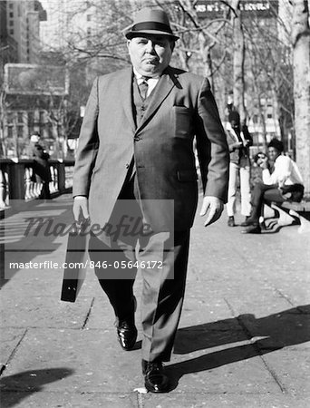 1960s OVERWEIGHT BUSINESSMAN WITH BRIEFCASE SMOKING CIGAR WALKING ON CITY SIDEWALK