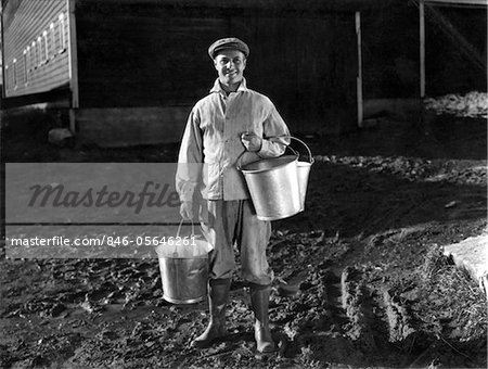 1930s - 1940s MAN FARMER DAIRYMAN STANDING OUTSIDE BARN HOLDING MILK PAILS WEARING CAP JACKET RUBBER KNEE BOOTS SMILING LOOKING AT CAMERA