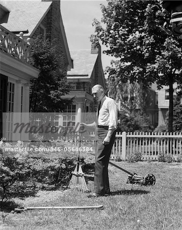 1930s SENIOR MAN STANDING BY GARDEN SHRUBS WITH RAKE PUSH MOWER LAWNMOWER BEHIND HIM