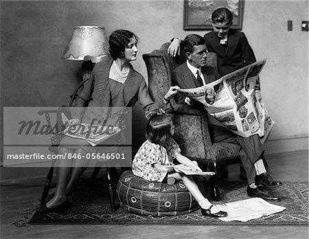 1920s FAMILY OF FOUR MOTHER FATHER SON DAUGHTER IN LIVING ROOM READING NEWSPAPER TOGETHER INDOOR