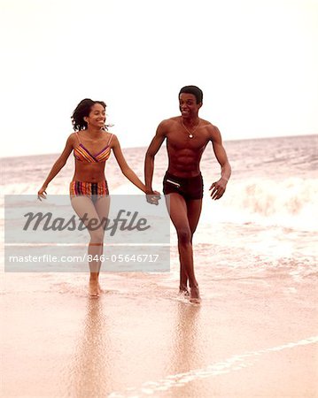 1970s AFRICAN AMERICAN  YOUNG COUPLE WEARING BATHING SUITS RUNNING IN OCEAN SURF