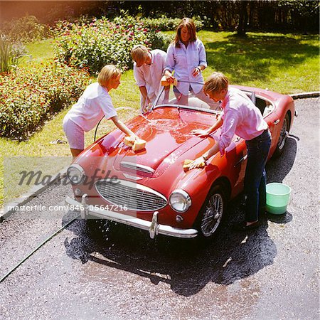 1970s FOUR TEENAGERS WASHING RED AUSTIN HEALEY SPORTS CONVERTIBLE AUTOMOBILE MAN WOMAN OVERHEAD OUTDOOR