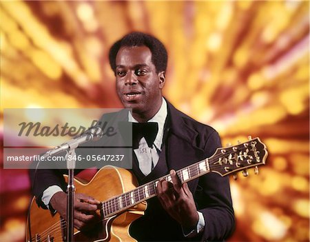 1970s AFRICAN AMERICAN MAN ENTERTAINER SINGER MUSICIAN WEARING TUXEDO PLAYING GUITAR SINGING INTO MICROPHONE GOLD LIGHTS BACKGROUND