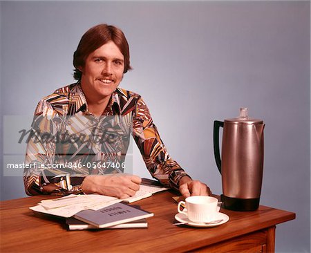 1970s YOUNG MAN MUSTACHE LOUD PRINT SHIRT DESK EMPTY COFFEE CUP POT WORKING ON FAMILY BUDGET LOOKING AT CAMERA