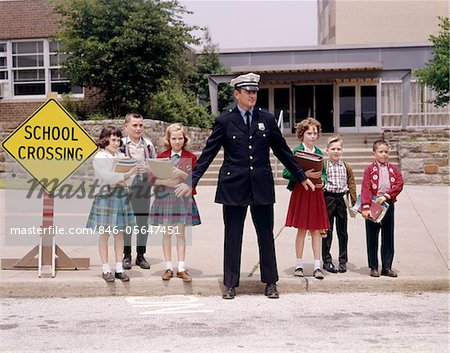 1960s MAN TRAFFIC POLICE OFFICER HOLDING BACK GROUP OF ELEMENTARY SCHOOL CHILDREN WAITING AT CURB TO CROSS STREET IN FRONT OF SCHOOL