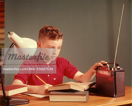 1960s RED HAIR BOY SITTING AT DESLISTENING TO PORTABLE RADIO WHILE STUDYING HOMEWORK 1960s RED HAIR BOY SITTING AT DESLISTENING TO PORTABLE RADIO WHILE STUDYING HOMEWORK